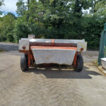 Orange agricultural trailer parked on a paved lot with green hedges and trees in the background; a green gate is on the right.