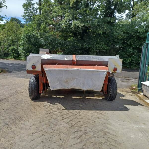 Orange agricultural trailer parked on a paved lot with green hedges and trees in the background; a green gate is on the right.