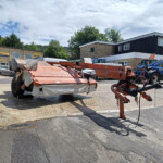 Industrial yard with orange agricultural machinery in the foreground and a beige brick building with New Holland signage in the background.