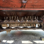 Rusty red agricultural implement with curved metal tines arranged in a row beneath the frame