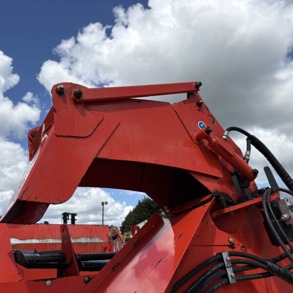 Red metal framework of heavy machinery with hydraulic hoses and bolts, set against a cloudy blue sky outside.