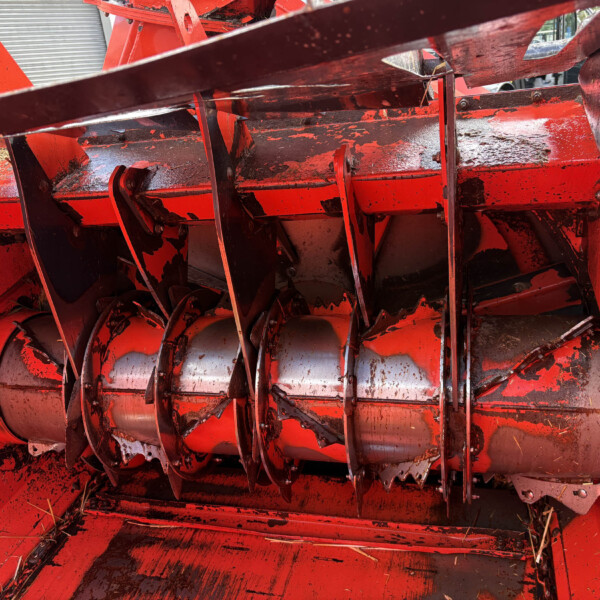 Close-up of a red farming machine's rotating cutting reel with metal teeth and chipped paint.