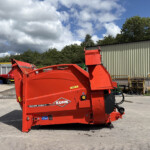 Bright red KUHN farm implement in an outdoor storage yard near a beige corrugated metal building, under a partly cloudy blue sky.