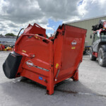 Red KUHN Primor 2060 M baler in an outdoor equipment yard beside a beige warehouse under a cloudy sky