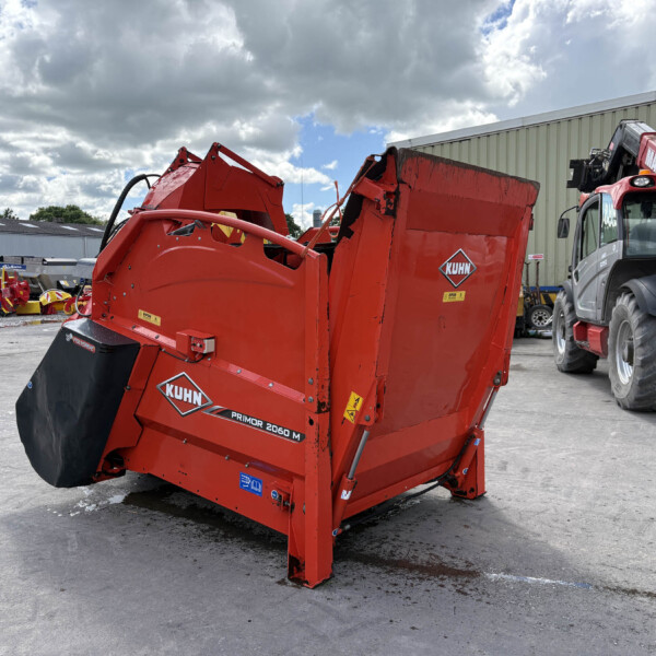 Red KUHN Primor 2060 M baler in an outdoor equipment yard beside a beige warehouse under a cloudy sky