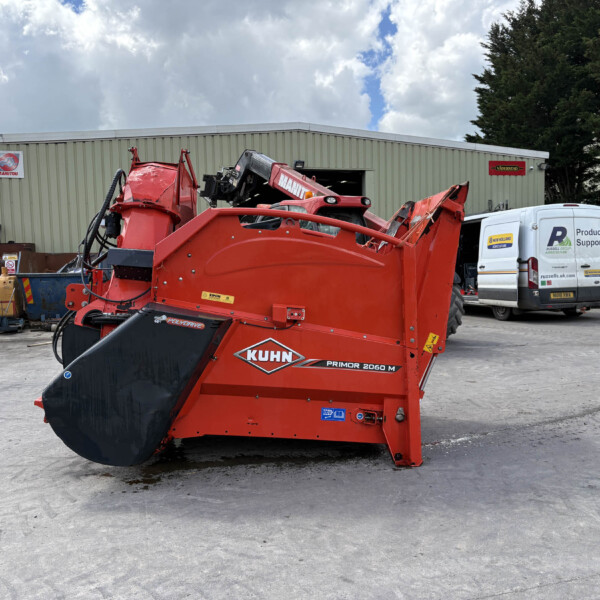Red industrial agricultural machine (KUHN Primor 2060 M) in a yard, with a gray building and a white van in the background.