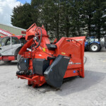 Industrial yard scene with red heavy machinery in the foreground and blue tractors parked by a warehouse under a cloudy sky.