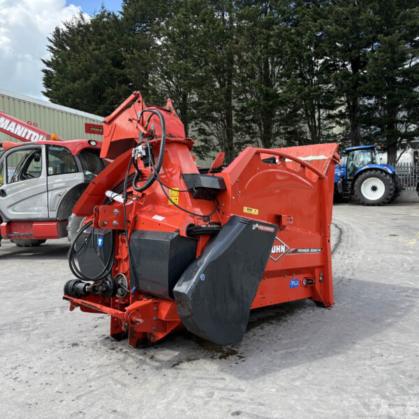 Industrial yard scene with red heavy machinery in the foreground and blue tractors parked by a warehouse under a cloudy sky.