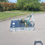 Metal trailer hitch with green straps on a paved road, a grassy curb and flowering hedge in the background.