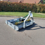 Trailer-mounted hydraulic lifting device on a residential street, secured with a green strap, with hedges and red-tiled houses behind.