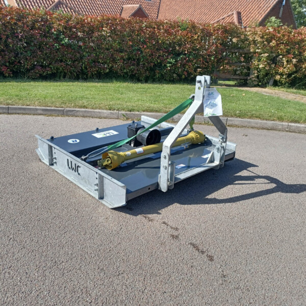 Trailer-mounted hydraulic lifting device on a residential street, secured with a green strap, with hedges and red-tiled houses behind.