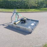Metal platform/trailer base on a paved surface, secured with green ratchet straps, with grass and bushes in the background.