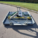 Rear view of a metal trailer hitch and winch on a flatbed trailer parked on asphalt, with grass and shrubbery in the background.