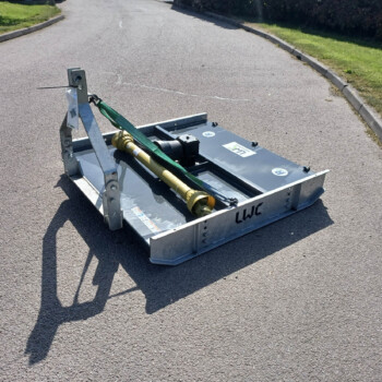 Close-up of a metal trailer hitch and hydraulic components secured with a green strap on an asphalt road with grassy verges nearby.