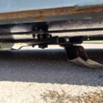 Underside view of a trailer hitch and metal frame under a trailer, resting on an asphalt surface with rusted components visible.