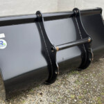 Black metal excavator bucket attachment resting on gravel beside a light concrete wall, outdoors with rust on edges and mounting pins visible, used for digging.