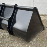 Large black steel excavator/skid steer bucket resting on gravel beside a white wall