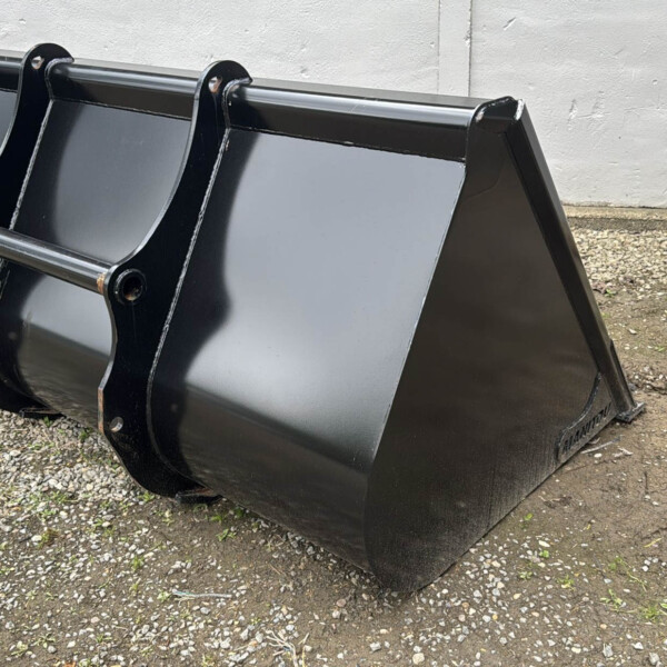 Large black steel excavator/skid steer bucket resting on gravel beside a white wall
