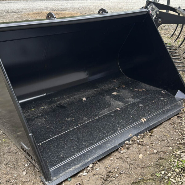 Heavy black steel bucket attachment resting on gravel outdoors, with skid-steer linkage visible at the top edge.