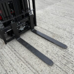 Close-up of black forklift forks extending over a textured concrete surface, with part of the forklift frame visible.