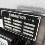 Metal nameplate on a rugged machine showing the Manitou logo and model/serial data, partially wet with raindrops.