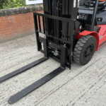 Red forklift with its black forks extended, parked beside a brick wall on a concrete surface, viewed from the front-left side.