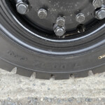 Close-up of five lug nuts securing a black wheel hub, with a dirty tire and gravel ground visible around it.