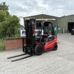 Red Manitou forklift with long forks resting on the ground in an industrial yard, in front of a green warehouse labeled 'RUSSELLS'.