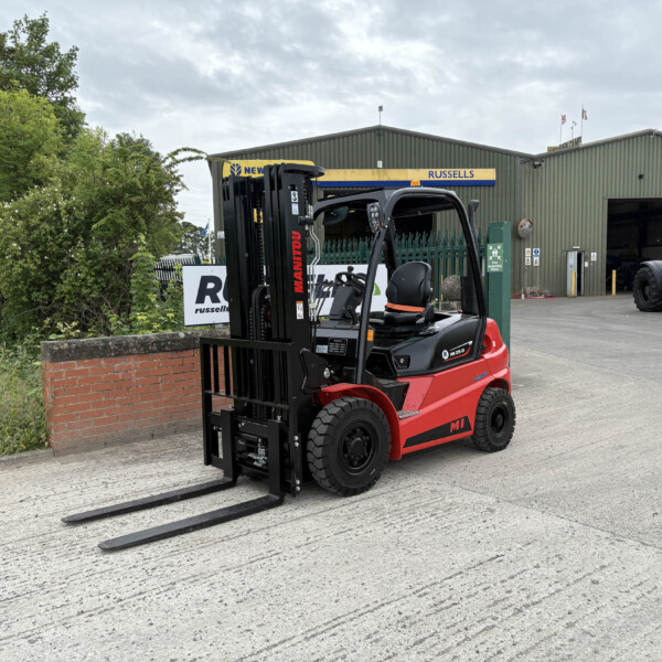 Red Manitou forklift with long forks resting on the ground in an industrial yard, in front of a green warehouse labeled 'RUSSELLS'.
