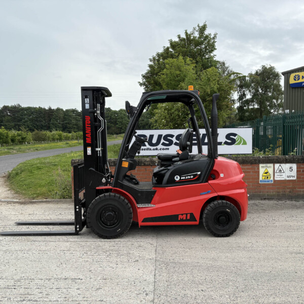 Red forklift with extended black forks parked on a paved lot near a brick wall and New Holland Agriculture building signage.