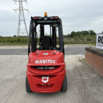 Front view of a red Manitou forklift with an orange beacon, outdoors beside a brick wall and highway.
