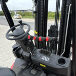 Close-up view of a forklift cab: steering wheel, multiple control levers with red handles, pedals, and dashboard, with an outdoor lot visible through the window behind.