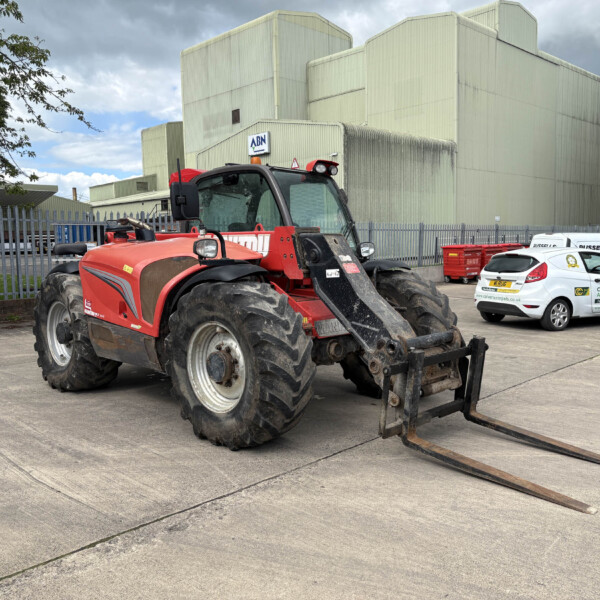 Red rough-terrain forklift with extended forks in a paved industrial yard, beige metal buildings in the background and a white van nearby.