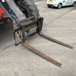 Close-up of rusted forklift forks resting on a concrete surface, with a large tractor wheel nearby and parked cars in the background.