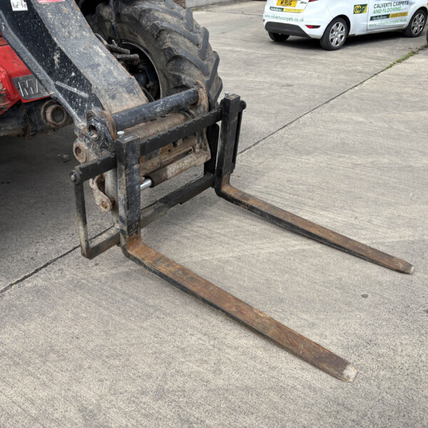 Close-up of rusted forklift forks resting on a concrete surface, with a large tractor wheel nearby and parked cars in the background.
