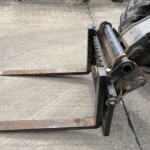 Rusty forklift forks extended forward over a worn concrete surface, with part of the hydraulic lift on the right side.