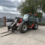 Red telescopic forklift with extended forks parked in an industrial parking lot by a metal fence and warehouse building on a cloudy day.
