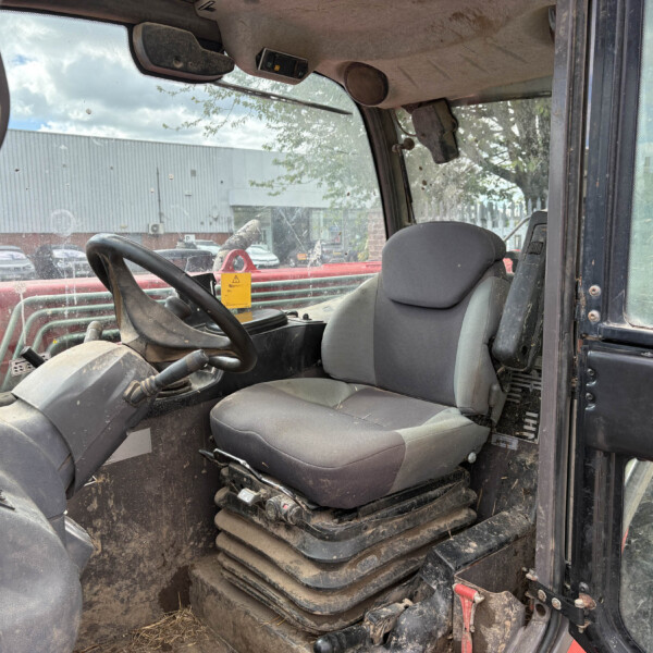 Interior of a dusty construction vehicle cab with steering wheel, worn seat, and dirty windows; outdoor yard beyond the door.
