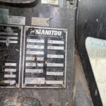 Close-up of a dirty metal identification plate on heavy equipment, showing the MANITOU branding and model/year labels.