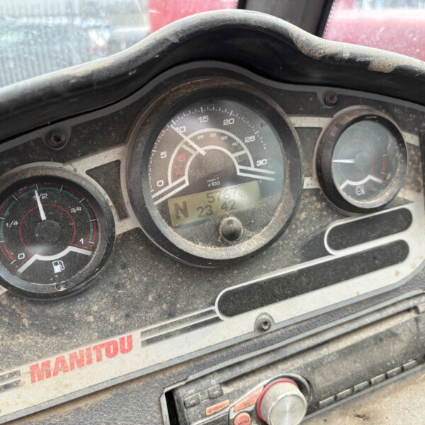 Dusty vehicle dashboard (Manitou) with round speedometer, fuel gauge, and control buttons visible behind a dusty windshield.