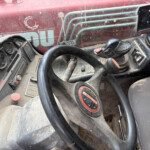 Interior of a dusty red tractor/cab showing the steering wheel, gauges, and control stalks across a dusty dashboard area with a red exterior visible through the window