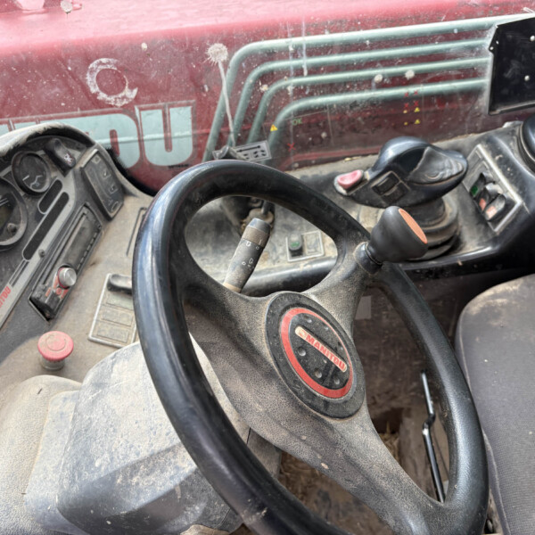 Interior of a dusty red tractor/cab showing the steering wheel, gauges, and control stalks across a dusty dashboard area with a red exterior visible through the window