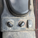 Close-up of dusty vehicle dashboard with two round knobs for fan and temperature and a snowflake defrost switch nearby.
