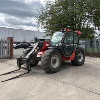 Red telescopic forklift with extended forks parked in an industrial parking lot by a metal fence and warehouse building on a cloudy day.