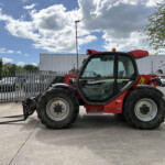 Red farm tractor with front loader forks lowered to the ground, parked in an industrial lot beside a metal fence and brick building under a blue sky with fluffy clouds.