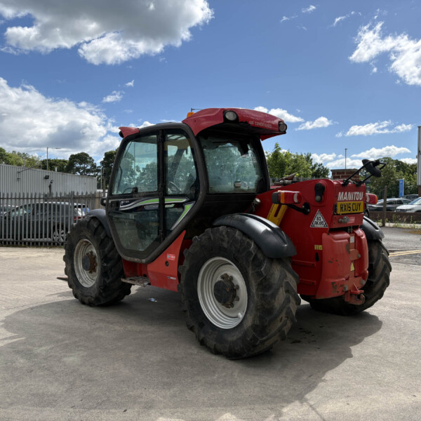 Red telehandler parked in an industrial yard with a metal fence and a blue sky with clouds