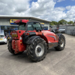 Red agricultural tractor in a parking lot outside a service building with a blue-framed window and cars nearby, under a cloudy sky.