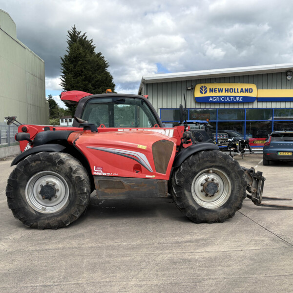 Red New Holland tractor in foreground at a dealership lot, with a blue-and-yellow New Holland Agriculture sign on a gray building and a cloudy sky above.