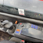 Interior of a heavy machinery cab with a large joystick, blue/orange buttons, and a dusty windshield