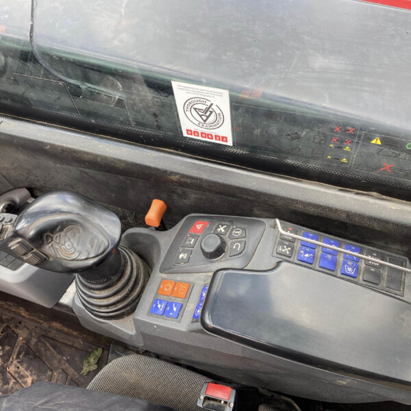Interior of a heavy machinery cab with a large joystick, blue/orange buttons, and a dusty windshield